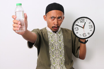 An Indonesian Muslim man in koko and peci holds a mineral water bottle and a clock, looking...