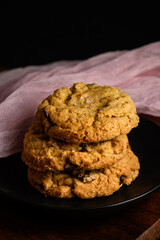 Spring treat, dark and moody image of three homemade fancy chocolate chip cookies stacked on a black plate with pale pink gauze clothe behind
