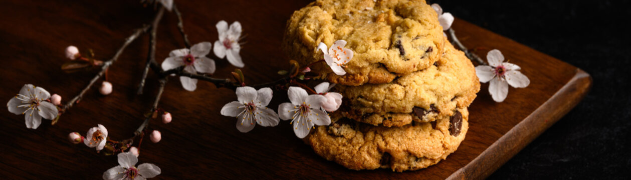 Spring treat, dark and moody image of three homemade fancy chocolate chip cookies stacked on a dark wood board with pale pink cherry blossom flowers
