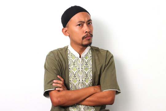 Thoughtful Indonesian Muslim man in koko shirt and peci crosses his arms, looking at the camera with a doubtful or confused expression. Isolated on White background