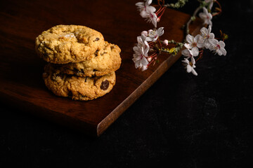 Spring treat, dark and moody image of three homemade fancy chocolate chip cookies stacked on a dark wood board with pale pink cherry blossom flowers
