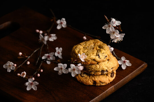 Spring treat, dark and moody image of three homemade fancy chocolate chip cookies stacked on a dark wood board with pale pink cherry blossom flowers
