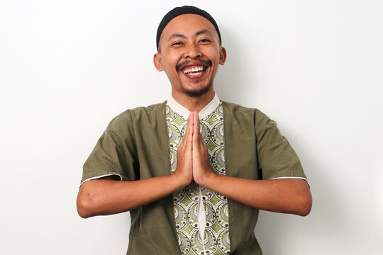 A happy Indonesian Muslim man in koko and peci smiles and gestures the traditional Eid Mubarak greeting, welcoming Ramadan. Isolated on a white background