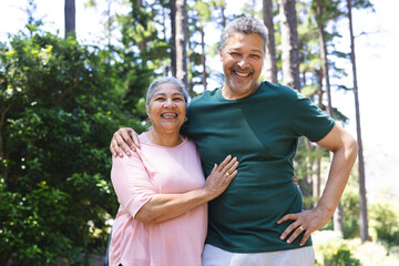 Senior biracial couple smiles warmly, standing outdoors with trees in the background