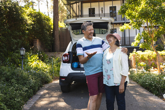 Senior biracial couple smiles while standing by their car in a sunny driveway