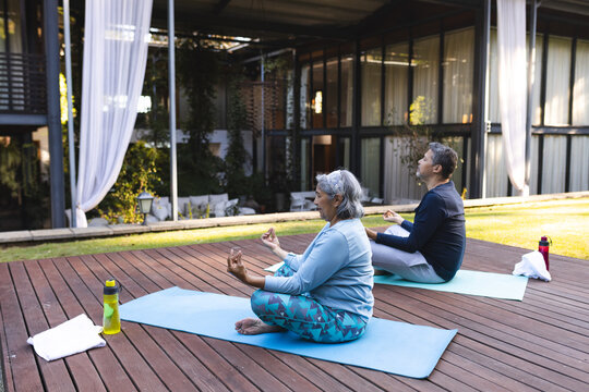 Senior biracial couple practices yoga outdoors on mats at home