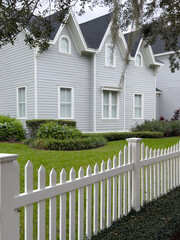A white wooden picket fence is in the foreground with lush green grass and hedges in the garden of...