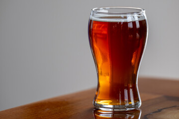 A small clear pint glass filled with a brewed red sour craft beer. The ale is on the corner of a wooden table. The top of the alcohol refreshment has white froth. The refreshing alcohol is a sample.