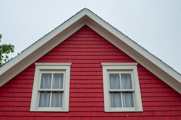 The middle roof peak of a vibrant red wooden cottage. The exterior wall of the house is covered in Cape Cod wood siding, two double-hung windows with cream trim and curtains. The sky is cloudy grey.
