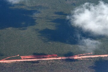 A straight highway runs through a forest. It is bordered by red dirt, with side roads of red dirt so that is looks like an arrow. The road is seen from the air, with clouds casting shadows.