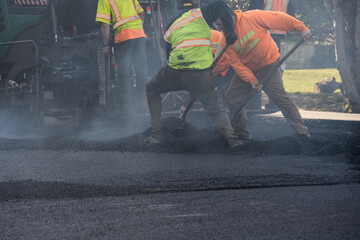 Residential street repaving project, workers in safety clothes shoveling fresh hot asphalt behind...