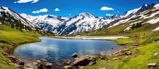 Obraz premium A mountain lake sits nestled among green grass and rugged rocks under the snowy peaks of Independence Pass. The tranquil water reflects the clear blue sky above.