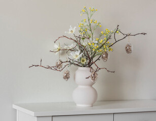 Easter bouquet of dry twigs, flowers, glass Easter eggs in a ceramic white vase on a white chest of drawers