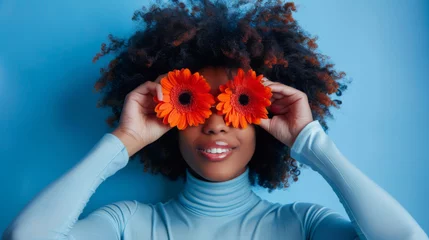 Plexiglas foto achterwand A person holds bright orange gerbera flowers over their eyes like glasses, smiling broadly against a blue background, creating a playful and joyful portrait. © MP Studio
