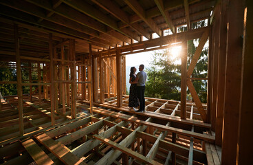 Man and woman inspecting their future wooden frame house nestled in the mountains near forest. Youthful couple at construction site in early morning. Concept of contemporary ecological construction.