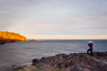 Une femme photographiant la côte méditerranée au lever de soleil. Lever de soleil sur le littoral. Touriste au bord de la mer en hiver. Lumière dorée sur la côte rocheuse.