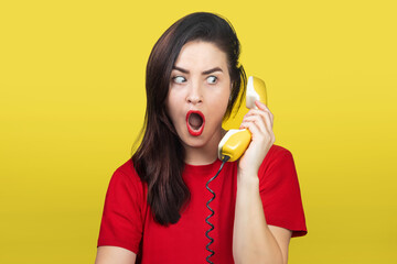 A beautiful brunette woman with red lipstick speaks on an old yellow telephone with a cord. She is shocked and surprised.