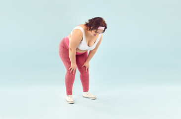 Fototapeta premium Portrait of a funny young fat overweight woman tired and resting after fit sport exercises wearing sportswear standing isolated on a studio blue background. Workout and body positive concept.