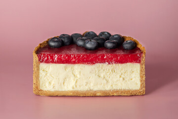 Half of vanilla cheesecake with berry confiture and blueberries on a pink background.