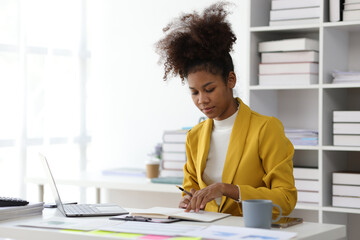 Attractive african american businesswoman working with laptop and documents on office desk writing work notes on notepad. - obrazy, fototapety, plakaty