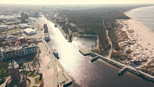 Drone captures sunny Kołobrzeg harbor in February, flying by lighthouse, cruise ships, and loading Ukrainian wheat onto cargo ship.