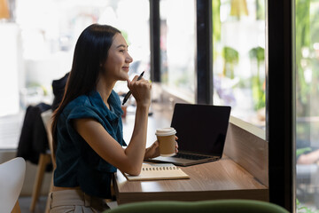 Contemplative young woman looking out window in coffee shop with laptop and coffee cup