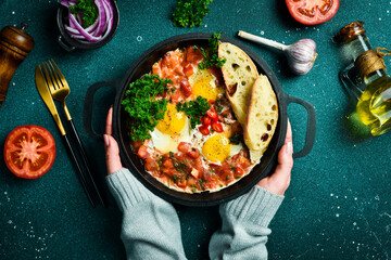 Hands hold a pan with Shakshuka eggs.. Poached eggs in a spicy tomato pepper sauce, onion and parsley. Top view. - obrazy, fototapety, plakaty