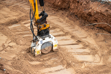A mini excavator rams the ground with a vibrating plate. Laying of underground sewer pipes and...