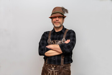 Studio portrait of a man wearing a Tyrolean feather hat and leather shorts, arms crossed on his chest, white background.