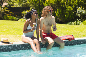 Diverse couple enjoys drinks by the poolside, laughter evident in their sunny disposition