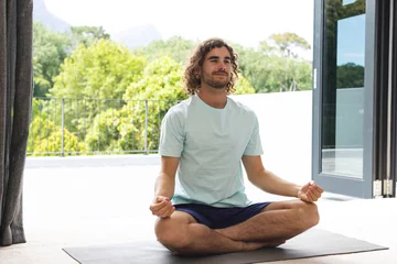 Poster Young Caucasian man with curly hair meditates in a lotus position on a yoga mat © wavebreak3