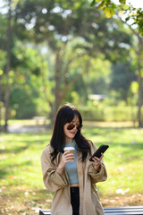 Portrait of young stylish woman in sunglasses drinking coffee and using mobile phone in the park