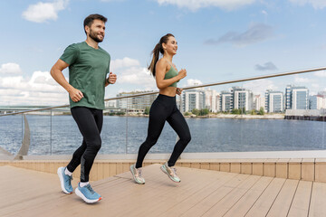 Fit couple running together on a wooden boardwalk by the river with urban skyline.