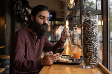 Male hipster eating healthy food sitting with smartphone in the loft cafe interior near window