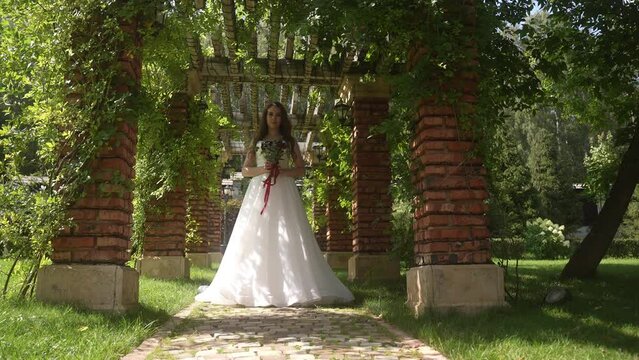 Bride in wedding gown under park archway, surrounded by trees and grass