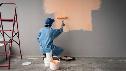 Painter with cap squatting painting a wall with paint roller, red ladder and buckets near. repair, building and home