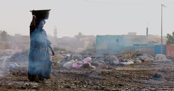 Close-up. Elderly black woman carrying fish in a plastic bucket on her head which have been smoked on wood chips. Air pollution. Dakar, Senegal