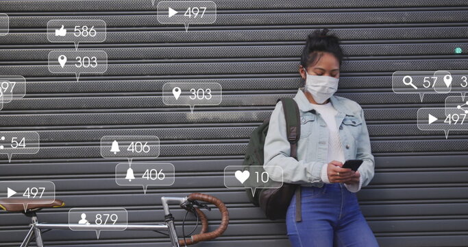 Image of social media icons and numbers on banners over woman in face mask using smartphone