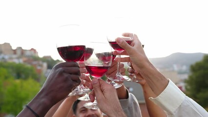 Group of young people raising wineglasses toasting red wine at rooftop summer party at sunset.