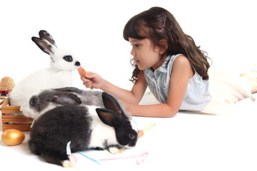 Cute little girl holds carrot to feed rabbit in wooden box. Joyful kid lies down on white background with golden easter eggs and pet, celebrating Easter day. Happy child on spring holiday celebration.