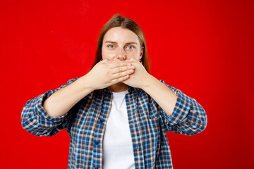 Young 30s woman covering closed mouth with hands open eyes. Speak no evil concept isolated red background. Human emotion facial expression concept
