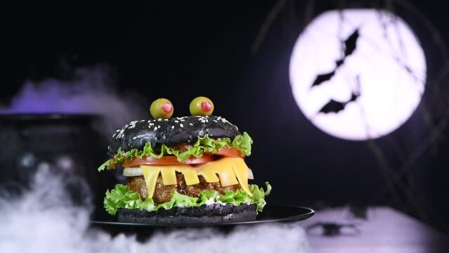 Monster Burger. Black bun, juicy beef cutlet, lettuce, onion, tomato and cheese in the shape of teeth, olive eyes. Halloween Party appetizer Idea. Slow motion, Circular rotation.