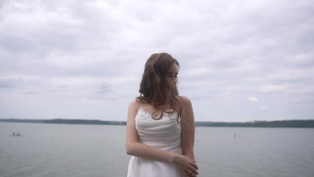 A bride stands at the lakeshore, flowers in hand, smiling under a cloudy sky