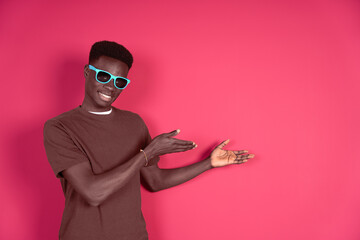 Young smiling black man wearing blue glasses with carmin red background pointing at the copyspace to put any text or virtual button isolated on red background.