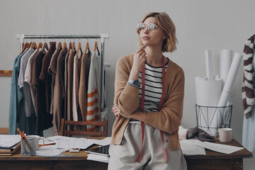 Attractive female fashion designer looking thoughtful while leaning at the desk in workshop - 749382965