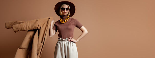 Fashionable young woman in elegant hat carrying her trendy coat against brown background - 749399533