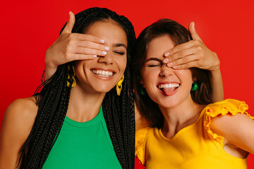 Two joyful young women in colorful wear covering eyes to each other and smiling against red background - 749409331