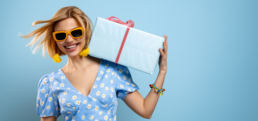 Beautiful young woman carrying a gift box on shoulder and smiling against blue background