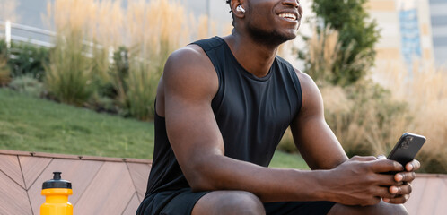 Handsome African man in sportswear using smart phone and smiling while having a break after training - 749414136