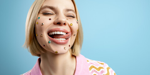 Happy young woman with colorful rhinestones over her face sticking out tongue against blue...